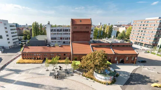 Aerial view of the EUREF Campus in Berlin with historic brick buildings and modern office complexes in the background. In the foreground, there are trees and a paved square