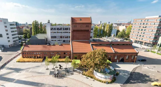 Aerial view of the EUREF Campus in Berlin with historic brick buildings and modern office complexes in the background. In the foreground, there are trees and a paved square
