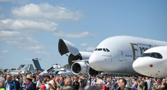 Large crowd at an airshow with airplanes, including an Emirates jet, on display under blue sky.