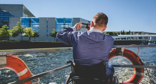 Man in a wheelchair taking a photo on a boat by a river, modern buildings in background.