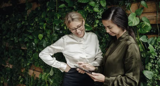 Two women in business attire stand in front of an ivy-covered wall and look at an Tablet with smiles on their faces.