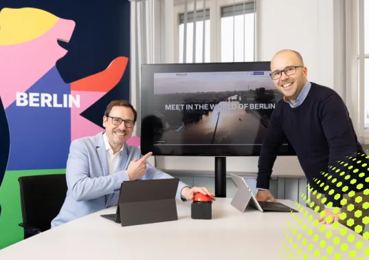 Zwei Männer lächeln an einem Tisch mit Laptops vor einem Bildschirm und Berlin-Poster.