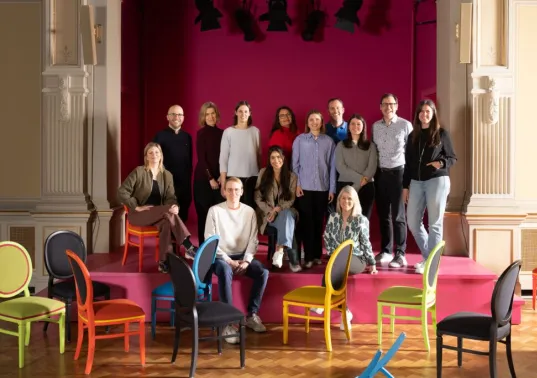 Group of people posing on a stage with colorful chairs in front, pink backdrop behind them.