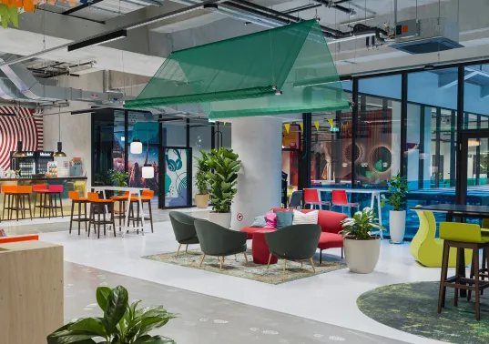 Modern office lounge with colorful furniture, plants, and large windows.