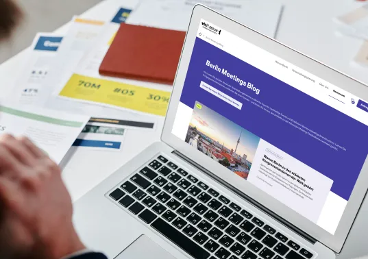 Person betrachtet Blogartikel auf Laptop-Bildschirm, Schreibtisch mit Unterlagen im Hintergrund.