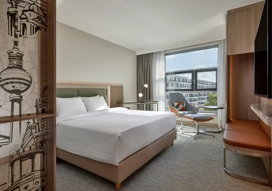Modern hotel room with a large bed, desk, chair, and window showing city buildings.