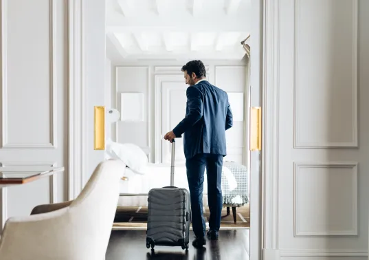 Mann im Anzug betritt mit Rollkoffer ein elegantes Hotelzimmer.