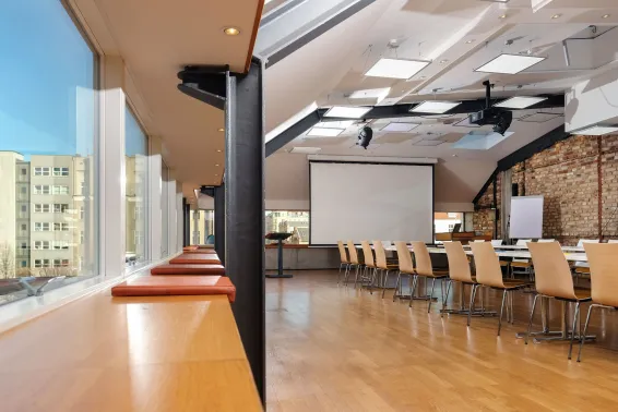 Heller Konferenzraum mit langen Tischen, Holzstühlen und großer Fensterfront