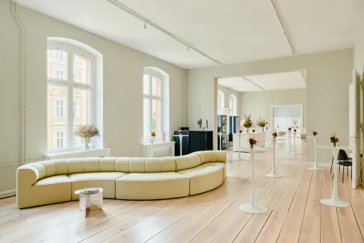 Heller & geräumiger Altbau-Saal mit Holzdielen-Boden, eingerichtet mit einem großes Design Sofa und Stehtischen mit kleiner Blumendekoration  