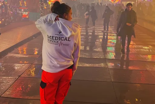 Sanitäterin mit weißer Jacke und roter Hose läuft nachts über einen bunten Jahrmarkt.