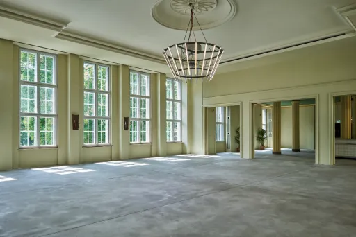 Großer, leerer Saal mit hohen Fenstern und Kronleuchter, viel Tageslicht.