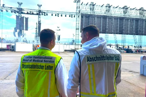 Zwei Sanitäter im weißen Outfit stehen vor einer großen Konzertbühne im Freien.