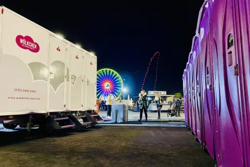 Hier sind auf der rechten Seite mehrere, pinke Wölkchen WC-Kabinen zu sehen. Auf der linken Seite steht ein mobiler WC-Wagen mit dem Logo, einer pinken Wolke und weißem Schriftzug "Wölkchen-Die frische Toilette". im Hintergrund steht ein sich drehendes Riesenrad.