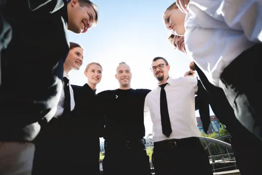 Sechs Menschen stehen lachend im Kreis, sonniger Tag, Teamstimmung.