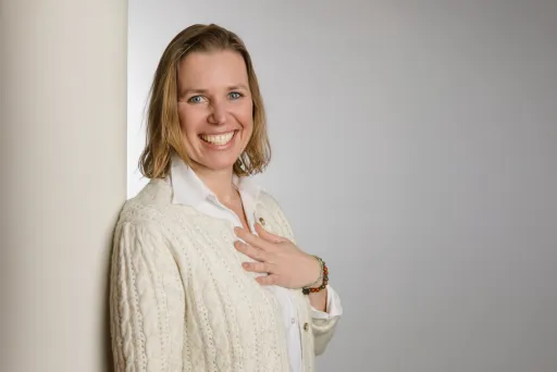 Frau mit hellem Pullover lächelt und lehnt an einer weißen Wand.