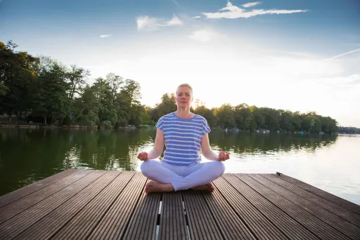 Person meditiert im Schneidersitz auf einem Steg am See bei Sonnenuntergang.