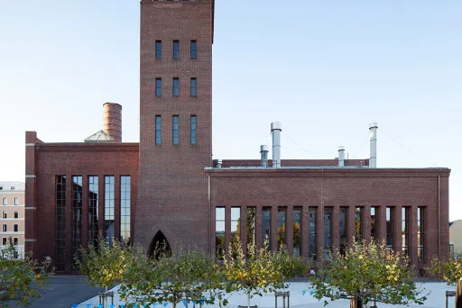 Backstein-Gebäude mit hohem Turm, davor Platz mit Bäumen und Sitzbänken.