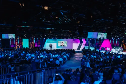 Großer Konferenzsaal mit vielen Zuschauern vor einer bunt beleuchteten Bühne.