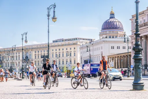 Mehrere Menschen fahren bei sonnigem Wetter Fahrrad vor historischen Gebäuden.