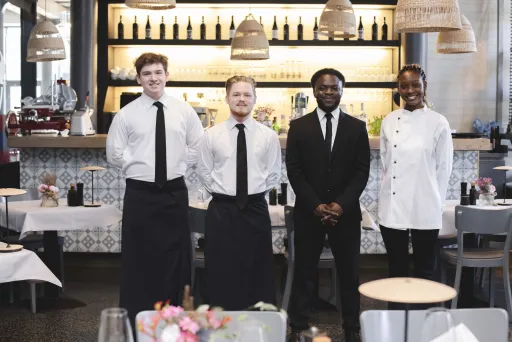 Vier Restaurantmitarbeiter stehen lächelnd vor der Bar im Gastraum.