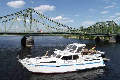 Weißes Motorboot auf Fluss vor grüner Stahlbrücke bei sonnigem Wetter.