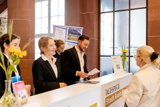 Mehrere Hosts und Hostessen in schwarzen Hosenanzügen stehen hinter der Akkreditierung und übergeben einer Dame einen Flyer – Eventpersonal und Promotionteam im Einsatz.