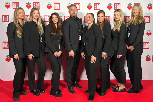 Acht Personen als Eventpersonal in schwarzen Hosenanzügen stehen auf rotem Teppich vor einer Wand mit den Logos ‚Ein Herz für Kinder‘ und ‚Bild Zeitung‘ – Promotion- und Eventteam im Einsatz.