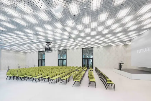 Moderner Konferenzsaal mit grünen Stühlen, großer Leinwand und futuristischer Decke.
