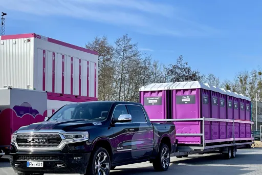 Hier wird ein Pick Up geuegt, der einen Anhänger zieht, auf dem mehrere, pinke Wölkchen WC-Kabinen stehen. Im Hintergrund ist ein WC-Wagen zu sehen, neben dem zwei, aufeinander stehende WC-Container, mit pinker Umrandung stehen.