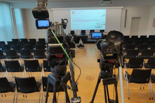 Leerer Konferenzraum mit zwei Kameras, Stuhlreihen und Bühne mit Projektionsleinwand.