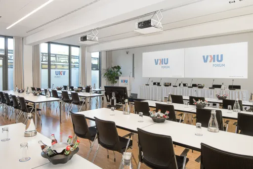 Heller Konferenzraum mit Tischen, Stühlen, Getränken und zwei großen Leinwänden.