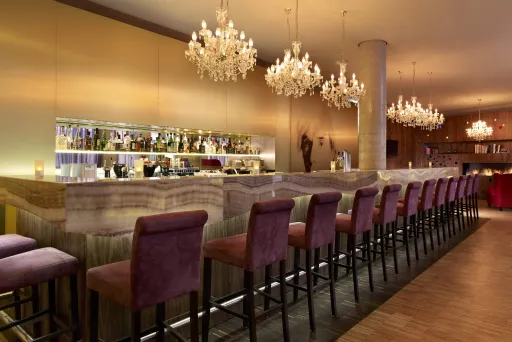 Moderne Bar mit lila Barhockern, beleuchteten Kronleuchtern und gut sortierten Regalen.