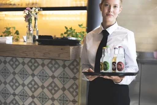 Kellnerin mit Tablett und drei Getränkeflaschen steht an einem Tresen.