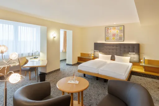 Helles Hotelzimmer mit Doppelbett, Sitzbereich und Schreibtisch, modern eingerichtet.