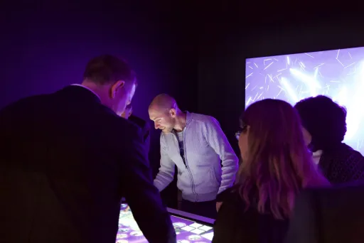 Mehrere Personen interagieren mit einem leuchtenden Touchscreen-Tisch in dunklem Raum.