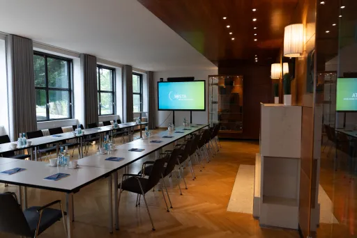 Moderner Konferenzraum mit langen Tischen, Stühlen und zwei Bildschirmen.