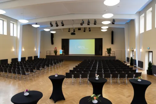 Heller Konferenzsaal mit Bühne, Stuhlreihen und Stehtischen, große Leinwand vorne.
