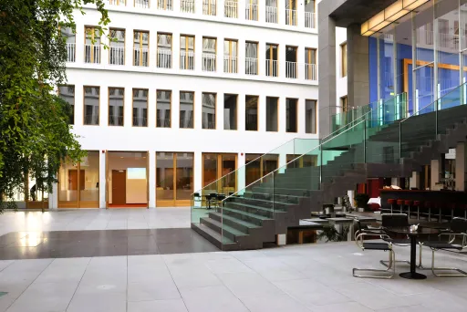 Modernes, helles Atrium mit Glastreppe und Tischen, große Fenster im Hintergrund.