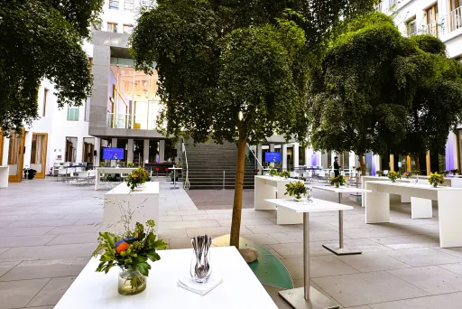 Moderner Innenhof mit weißen Tischen, Blumendekorationen und großen grünen Bäumen.