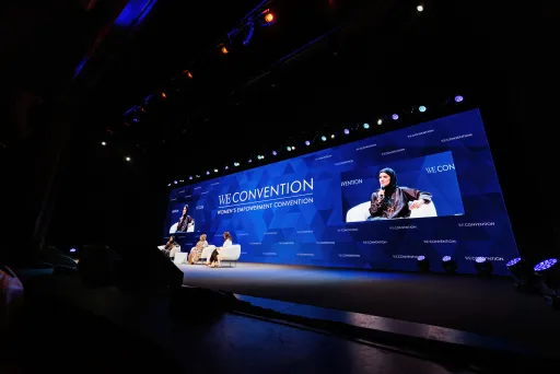 Das Bild ist dunkel. Zu sehen ist rechts im Hintergrund eine LED Wand, welche einen blauen Hintergrund zeigt, weiße Schrift drauf hat und zwei Bilder im Bild. Auf den Bildern zu sehen ist eine der drei Sprecherinnen, die vor der LED-Wand auf Sesseln sitzen.