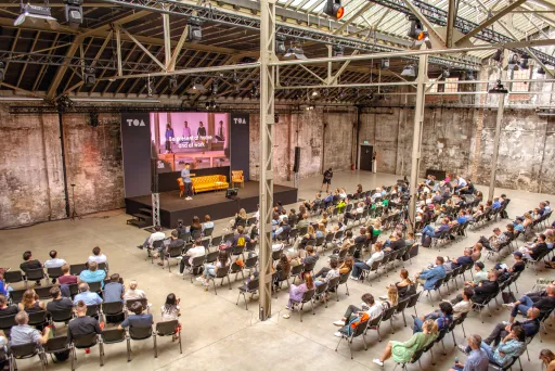 Zu sehen ist eine Fabrikhalle. Links gehalten im Hintergrund ist eine kleine Bühne, mit LED Wand, einem Sofa , ein Stuhl und einer Person, die darauf stehen.  Vor der Bühne sind Stühle in einigen Reihen aufgestellt und sie sind fast alle voll besetzt.