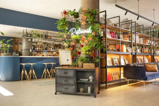 Modernes Café mit Pflanzen, Regal voller Bücher und gemütlicher Sitzecke.