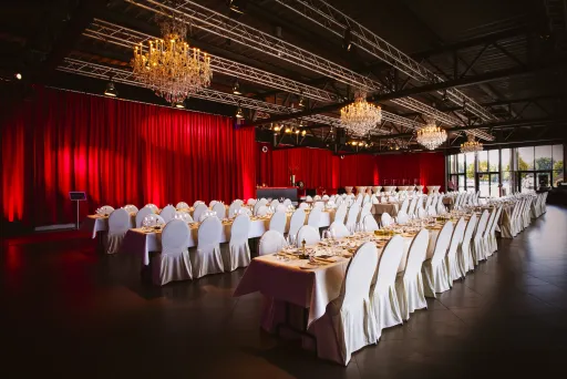 Eleganter Bankettsaal mit runden Tischen, weißen Stühlen und roten Vorhängen.