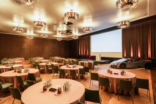 Konferenzraum mit runden Tischen, großer Leinwand und einem silbernen Auto auf der Bühne.