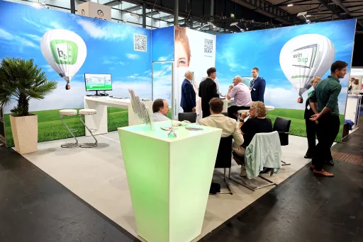 Messestand mit Menschen, grüner Theke, Stühlen und Himmel-Ballon-Motiv im Hintergrund.