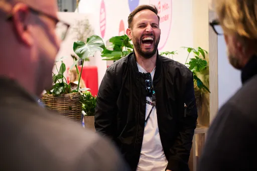 Mann lacht herzlich im Gespräch mit zwei anderen Personen, Pflanzen im Hintergrund.