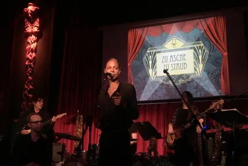 Sänger und Band auf Bühne mit rotem Vorhang und beleuchtetem "So Fackin Wild"-Schild im Hintergrund