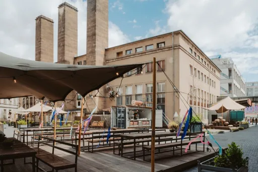 Städtischer Innenhof mit Holzbänken, Sonnensegeln und Backsteingebäuden im Hintergrund.