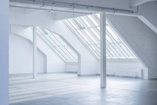 Großer, leerer Raum mit schrägen Fenstern und viel Tageslicht