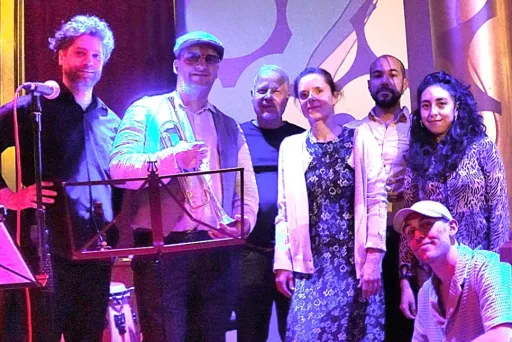 Gruppe von sieben Menschen auf einer Bühne, bunt beleuchtet, zwei halten Trommeln.